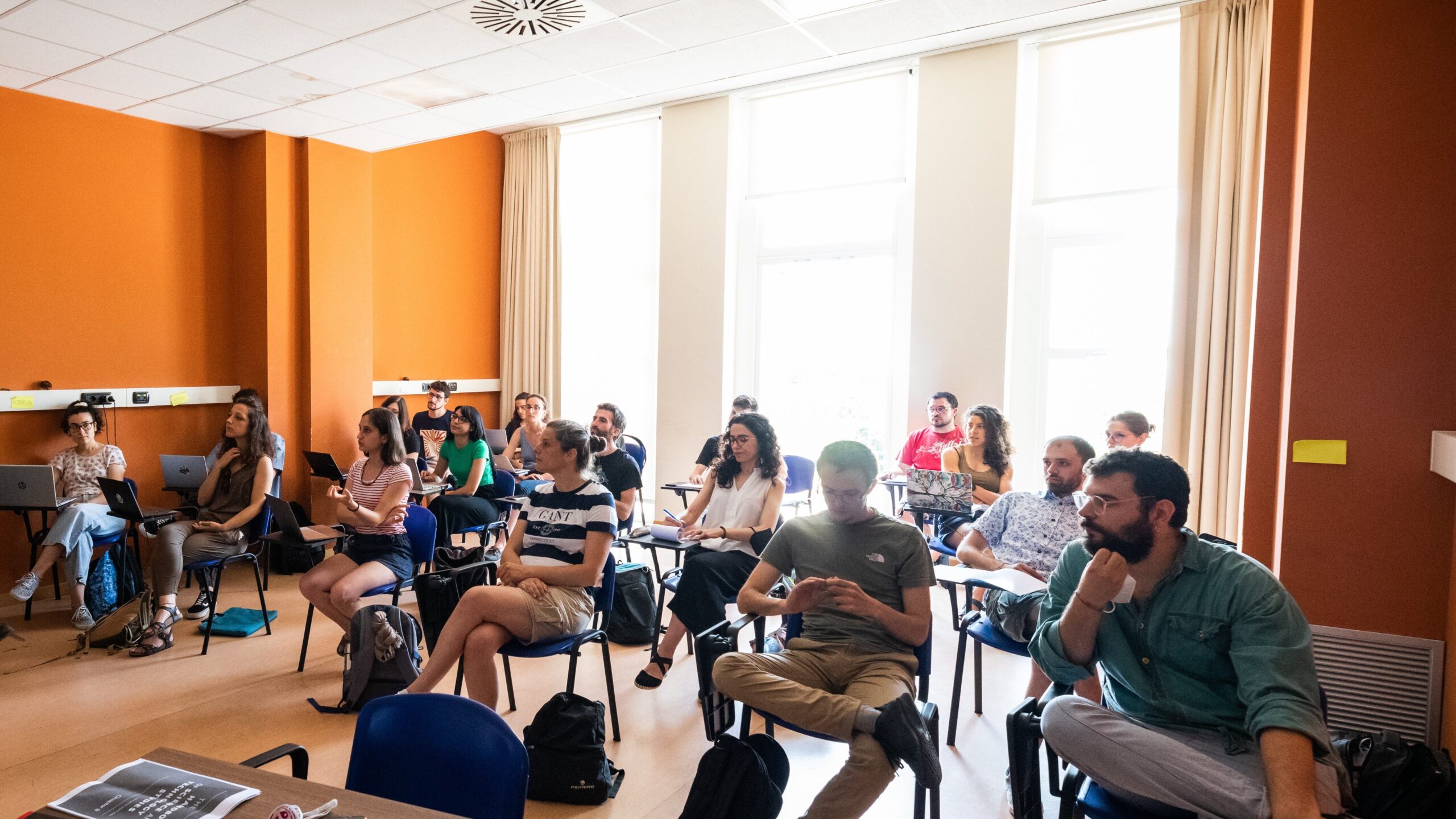 Lezione del Master in Comunicazione della Scienza Partecipanti seduti in aula durante una lezione del Master in Comunicazione della Scienza, con pareti arancioni e finestre lu
